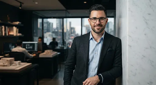Portrait of a male architectural consultant with circular glasses and a dark grey blazer leaning against a white marble pillar.